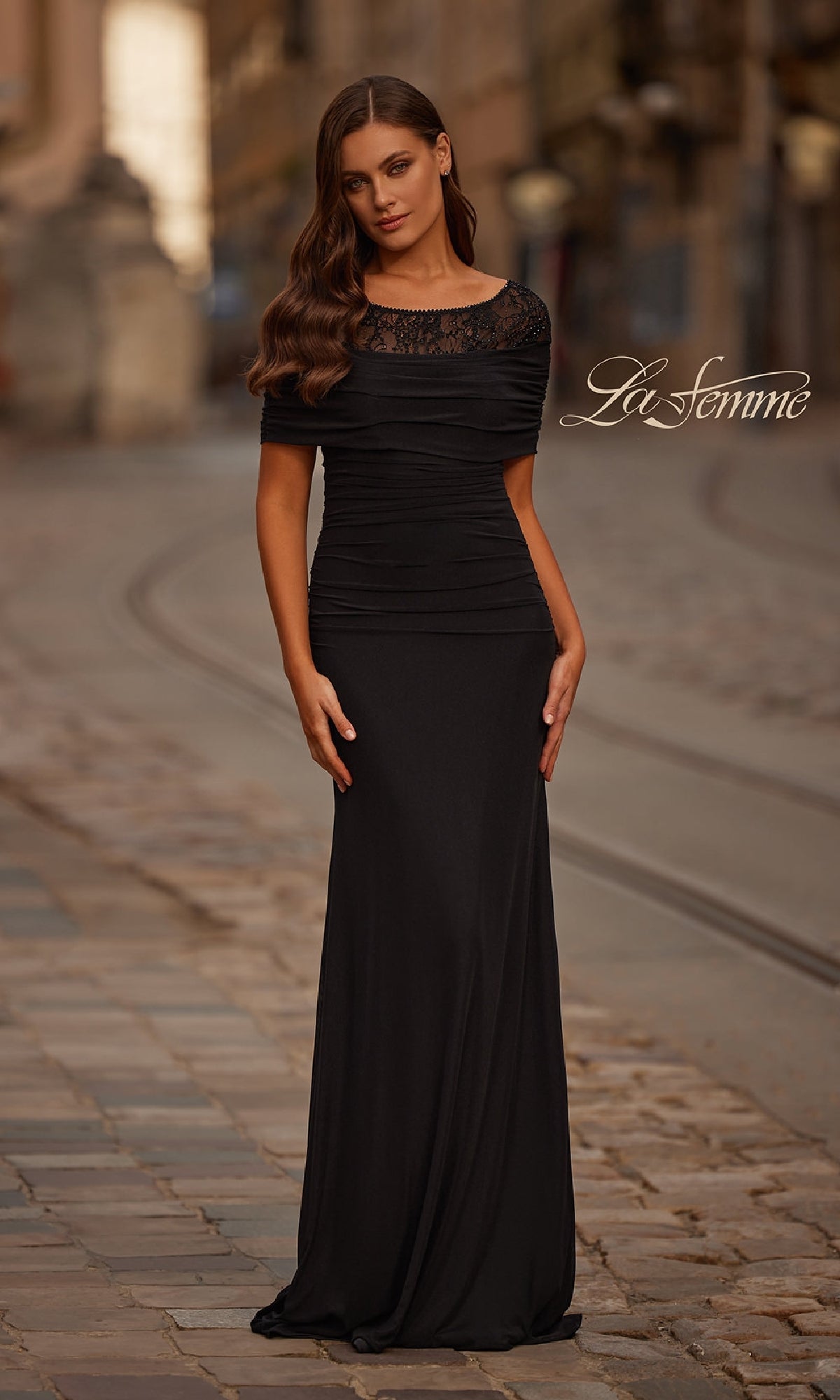 A woman in the Short Sleeve Black Formal Gown: La Femme 33120, detailed with beaded lace, stands on a cobblestone street; the